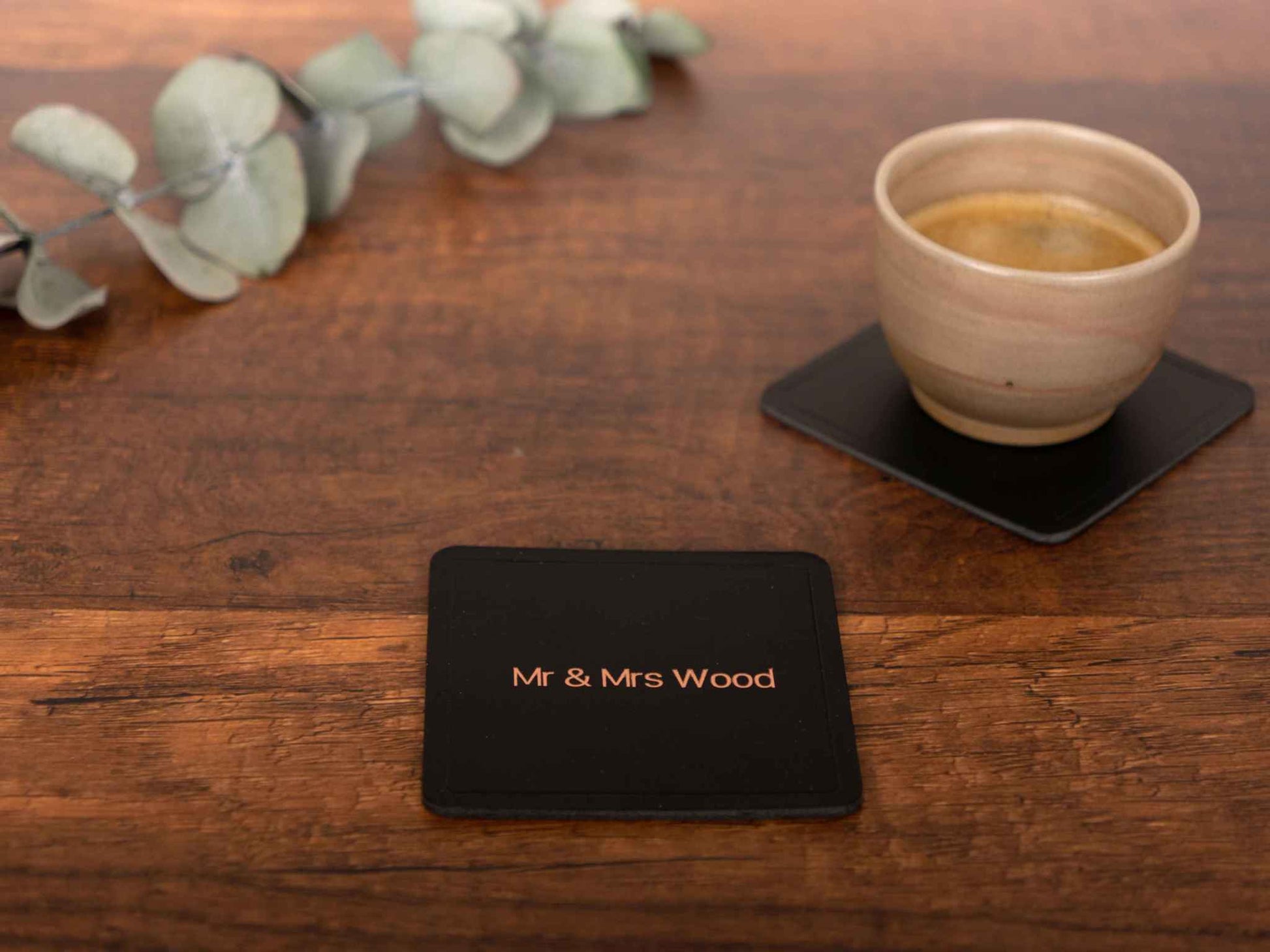 Gepersonaliseerde zwarte leren onderzetter met koperen folie-gravure "Mr & Mrs Wood" op houten tafel met koffiekopje op soortgelijke onderzetter