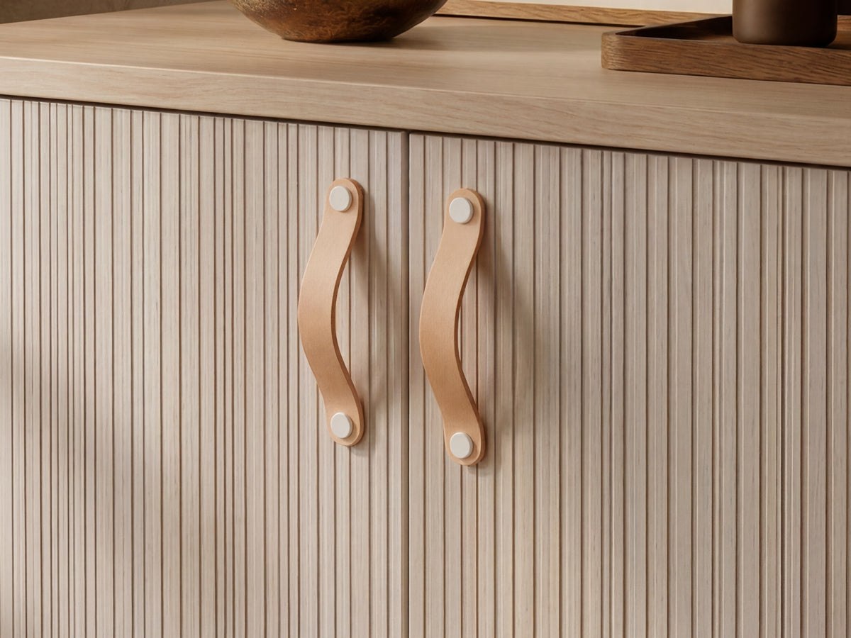 Close-up of a light wood cabinet with vertically fluted doors and two natural leather handles with white rivets, set against a matching wood countertop.