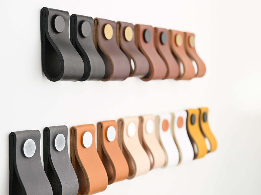 Assorted leather drawer pulls in various colors with metal fittings displayed in two rows on a white surface