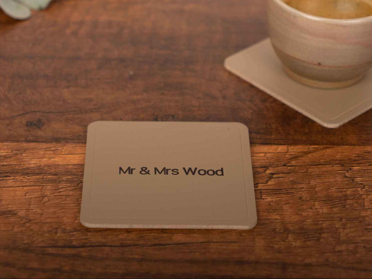 Sous-verre carré en cuir personnalisé avec gravure « Mr & Mrs Wood » sur une table en bois à côté d'une tasse à café.