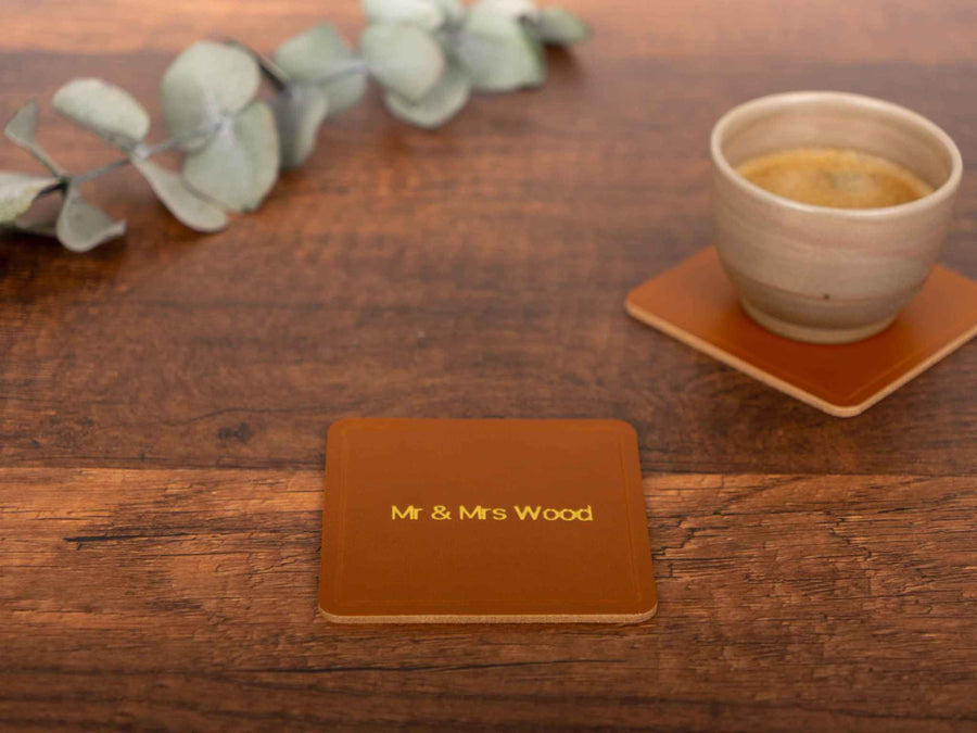Square personalized leather coaster in premium Italian leather with gold foil stamping on rustic wooden surface next to ceramic cup